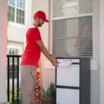A postal worker in a red uniform and cap places a letter into an outdoor package delivery box