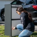 A woman wearing a black sweatshirt and jeans crouches to open a home package delivery box with her personal key