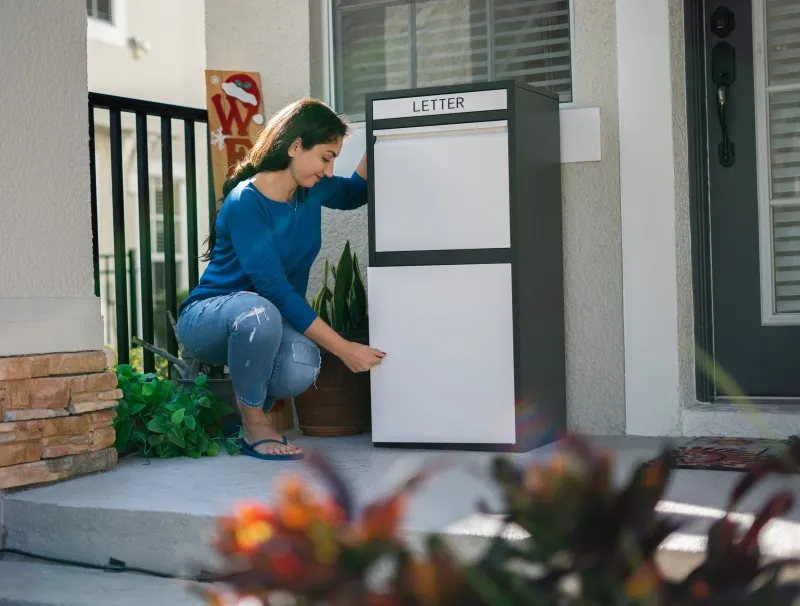 Una mujer con un suéter azul y jeans se agacha para colocar una carta en un gran buzón seguro para paquetes con la etiqueta "LETTER" situado en el porche de una casa.