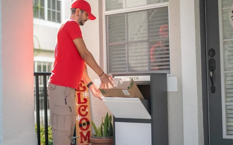 A delivery man in red is placing a large parcel into the white package delivery box located beside the homeowner's door.