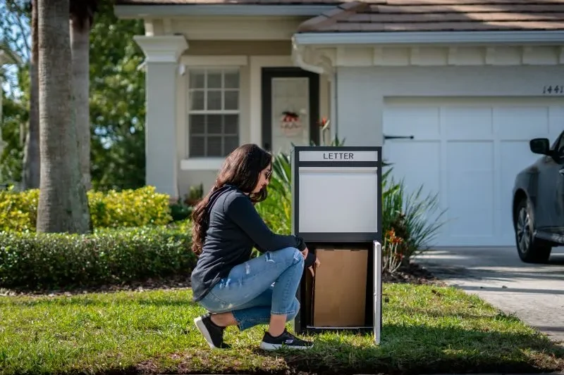 A woman dressed in black crouches to retrieve a package from a large white delivery box labeled "LETTER" in the front yard of a house.