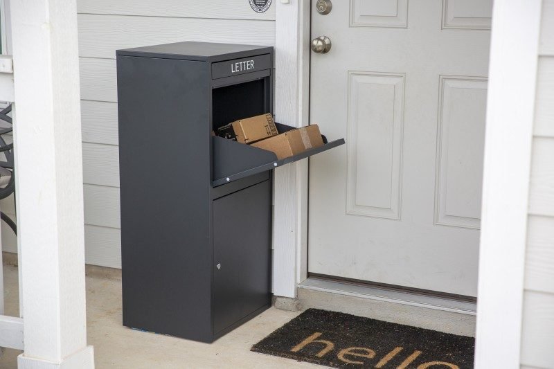 A black package delivery box is placed beside the door, with the top door open, revealing two parcels inside.